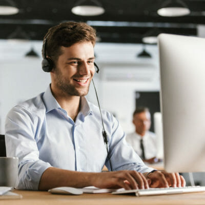 Photo of businesslike man 20s wearing office clothes and headset working on computer in call center